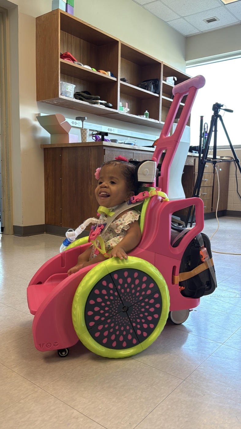 Side view of the Toddler Mobility Trainer in pink and lime green, with a laughing toddler aboard